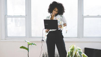 Woman standing and holding laptop with windows and plants in the background